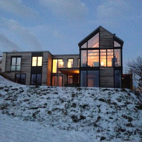 Et stort moderne hus midt i vinteren med masse store vindu og varm belysning sett fra utsiden